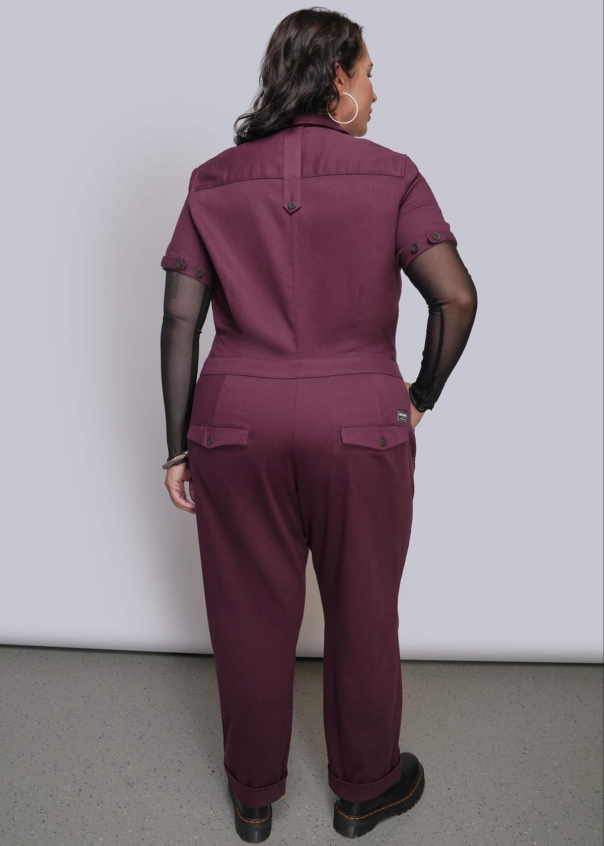 1X=A woman is shown from the back wearing a burgundy jumpsuit with a sheer black long sleeve top underneath, standing against a plain light gray studio background.