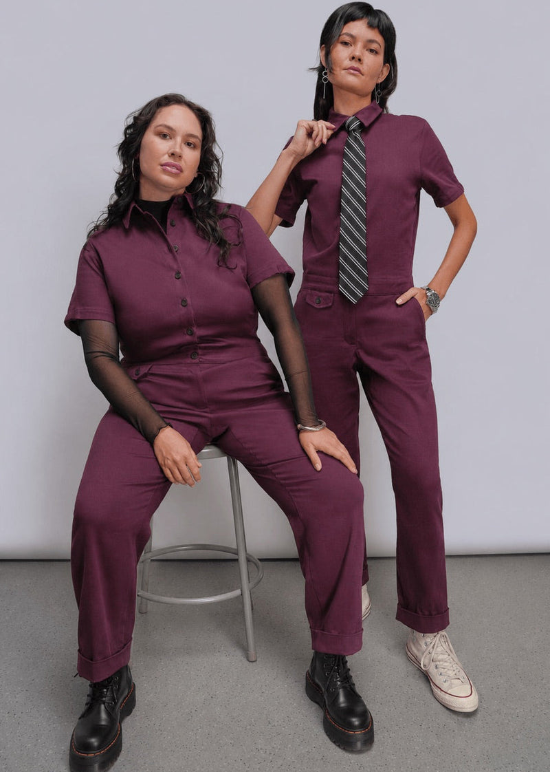 Two women modeling matching burgundy jumpsuits in a studio setting. One woman sits on a metal stool wearing a short-sleeve button-front scrub top and matching pants, while the other stands wearing the same uniform with a striped tie and white sneakers.