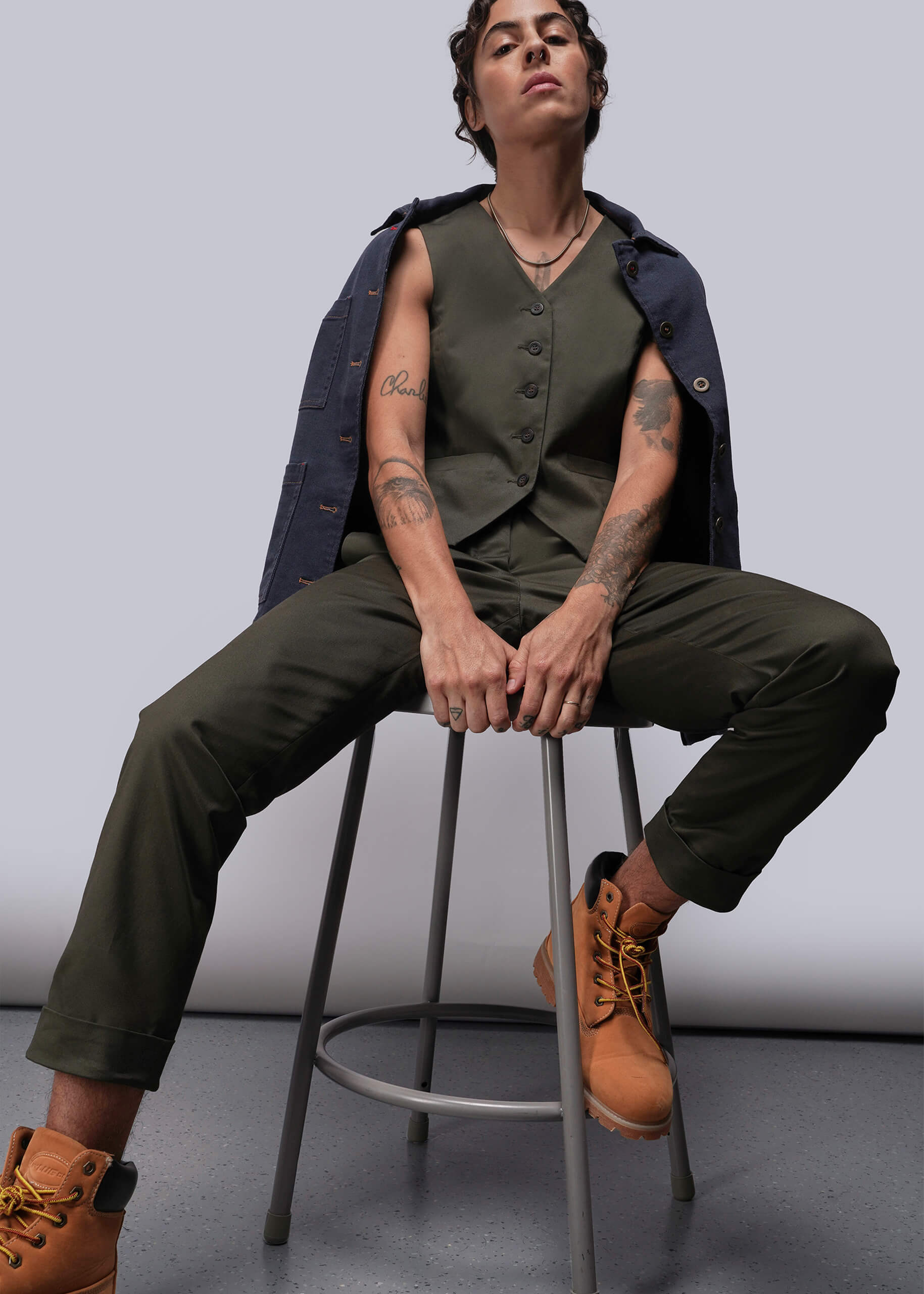 Person wearing a green vest and pants with brown boots sitting on a stool against a plain background