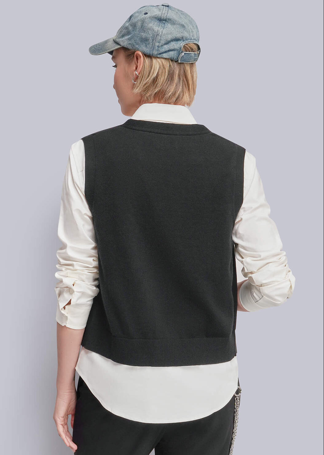 S=A model shown from the back wearing a black knit vest over a cream shirt, black trousers, and a blue denim cap against a light gray studio background.