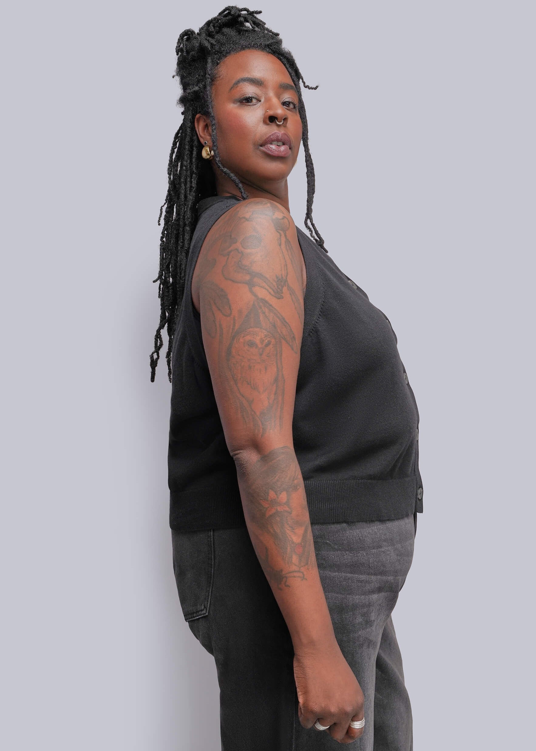 1X=A person with long black hair stands in profile wearing a black sleeveless top and dark gray jeans against a solid light gray studio background.