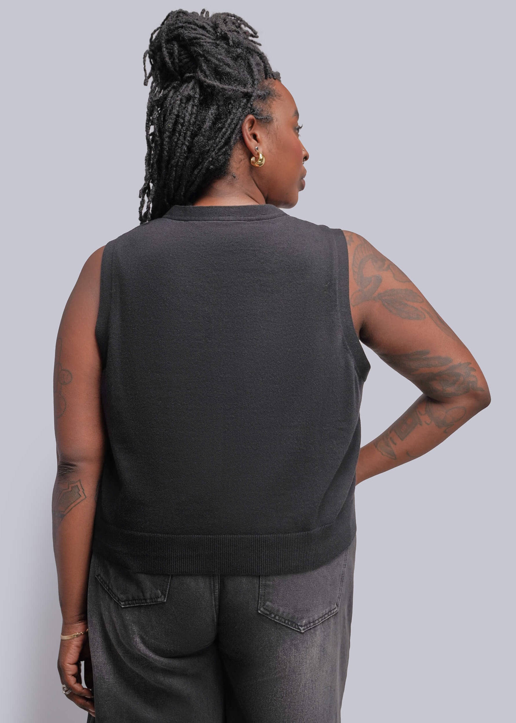 1X=A person shown from the back wearing a black gray sleeveless top and gray jeans against a light gray studio background.