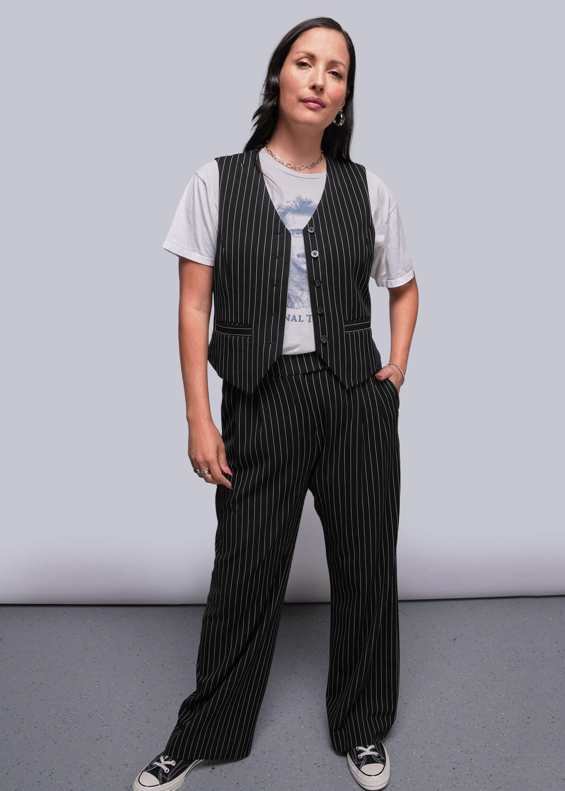 Model stands in front of gray background wearing pinstripe vest over t shirt and trouser 