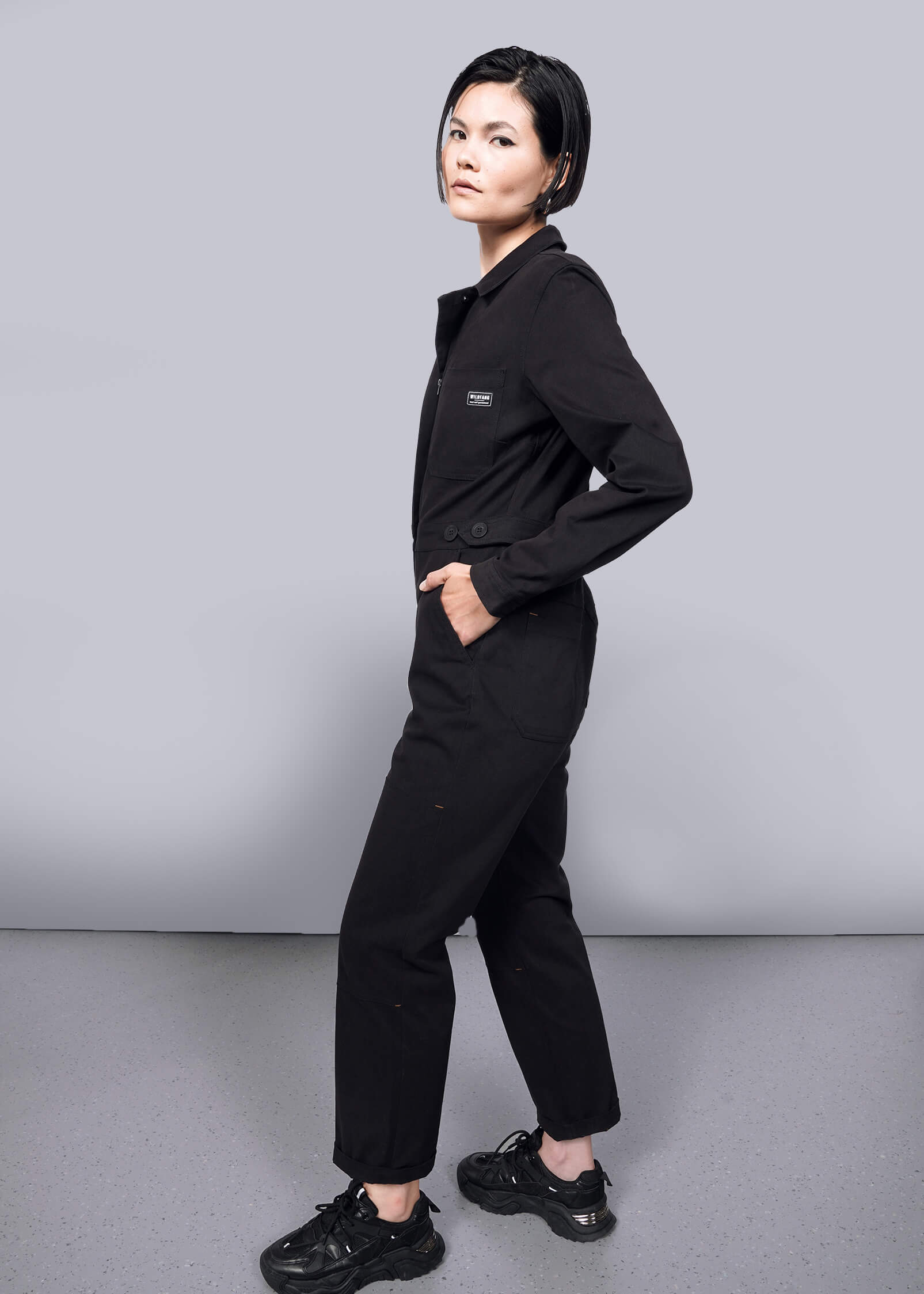 S=A person with short dark hair stands sideways, looking at the camera. They wear The Essential Long Sleeve High Waisted Coverall and black sneakers, posing against a plain white background and gray floor.