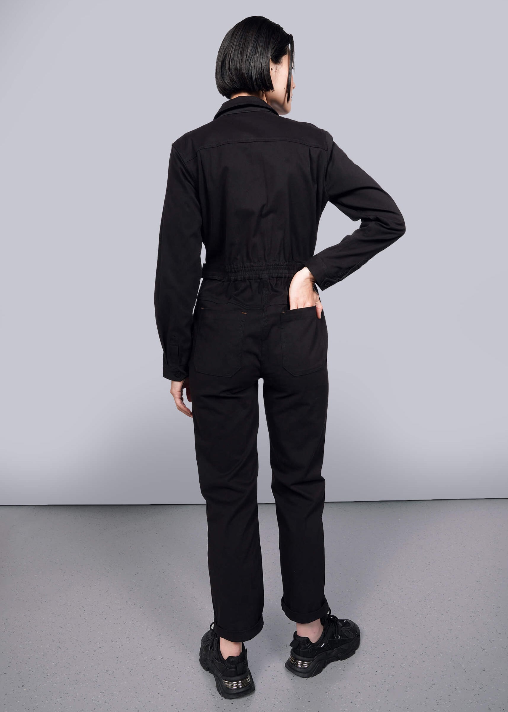 S=A person with short dark hair faces away, wearing The Essential Long Sleeve High Waisted Coverall and black sneakers, one hand on their hip and the other in a back pocket, set against a plain white background.