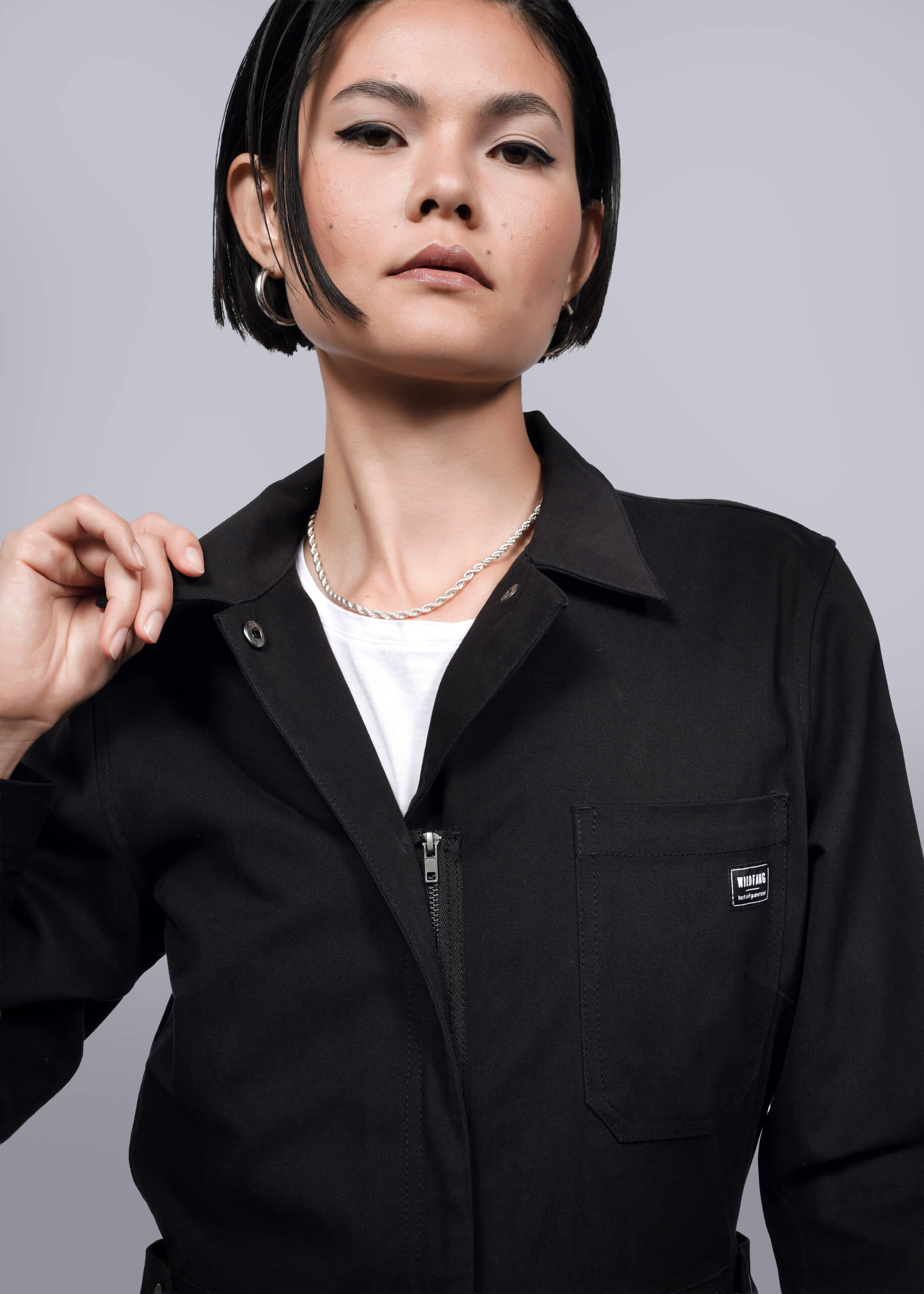 S=A person with short black hair wears The Essential Long Sleeve High Waisted Coverall in black over a white top, holding the collar. The background is plain white.