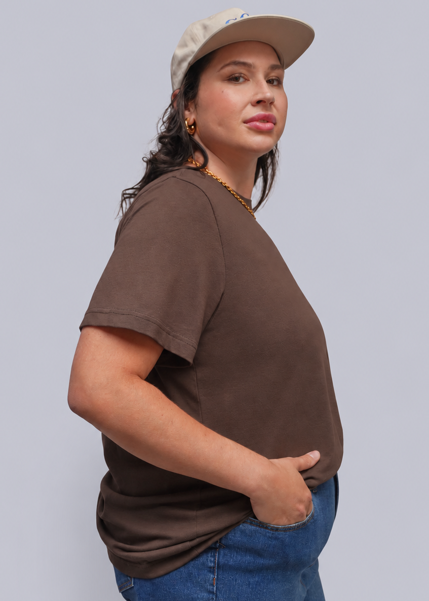 1X=A woman wearing a brown short-sleeve crewneck t-shirt, blue jeans, a gold chain necklace, and a beige baseball cap, shown from a side profile against a solid gray background.