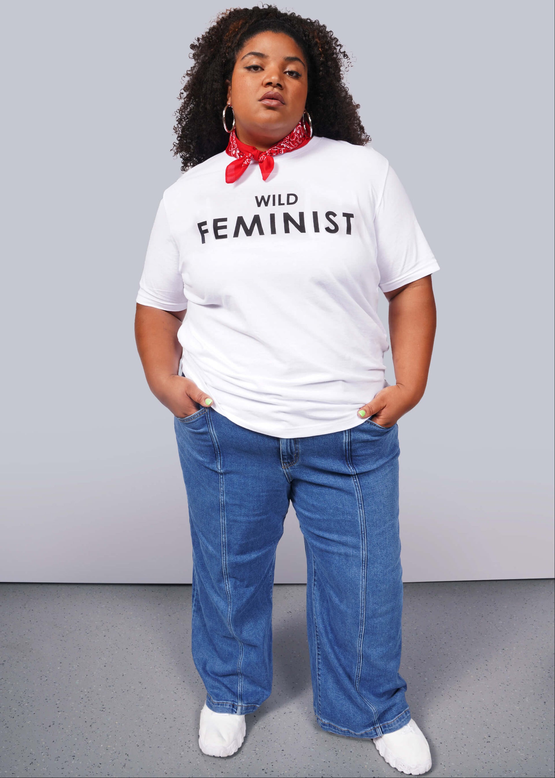 1X= A confident person wears The Wild Feminist® Tee, a relaxed unisex fit white shirt, paired with a red bandana, blue jeans, and white shoes. The plain background emphasizes their commitment to unity and advocacy.