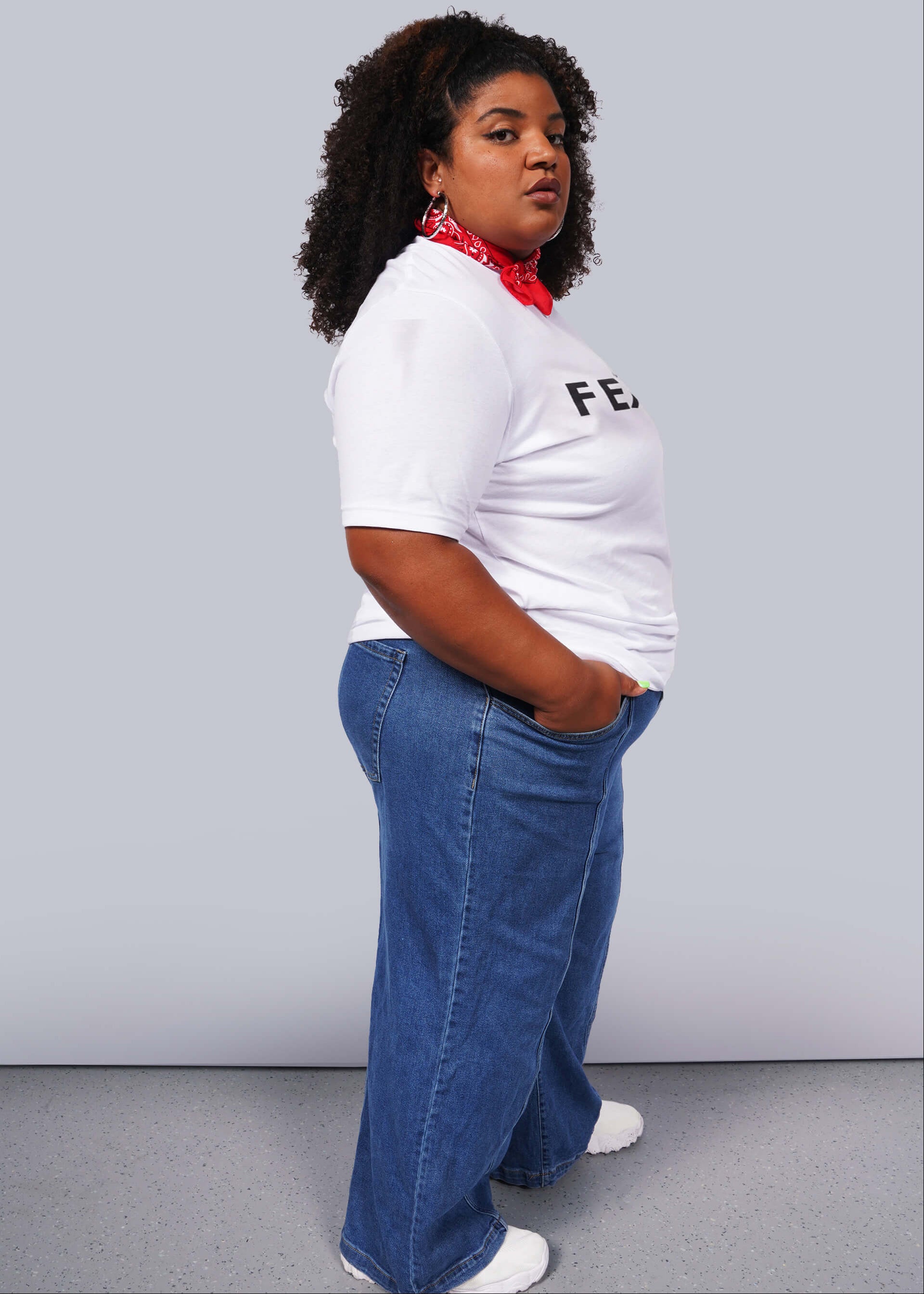 1X=A person with curly hair confidently poses against a gray background, wearing a relaxed unisex fit. They sport The Wild Feminist® Tee, blue jeans, white sneakers, and a red bandana around their neck. One hand in their pocket, they embody unity.