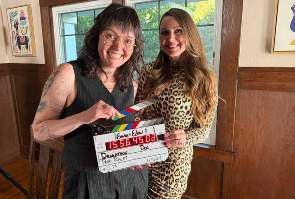 Two people holding movie clapper in front smiling at camera while standing inside house with wooden walls.