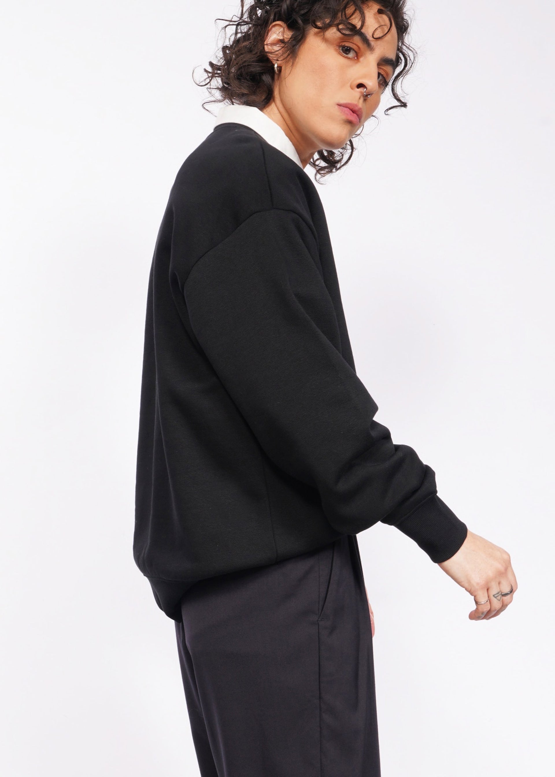 S=Someone with curly hair, sporting the Wild Feminist Fleece Crew in black and dark pants, poses against a light backdrop. Their expression is neutral, gazing slightly to the side.