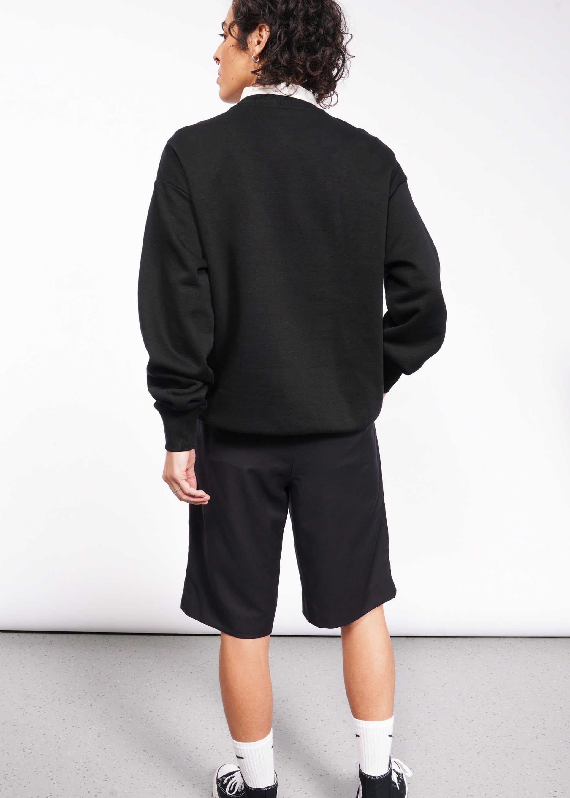 S=A person with curly hair stands against a plain background, dressed in a black Wild Feminist Fleece Crew, black shorts, white socks, and black sneakers. The brushed fleece adds comfort while subtly capturing the spirit of a WILD FEMINIST.