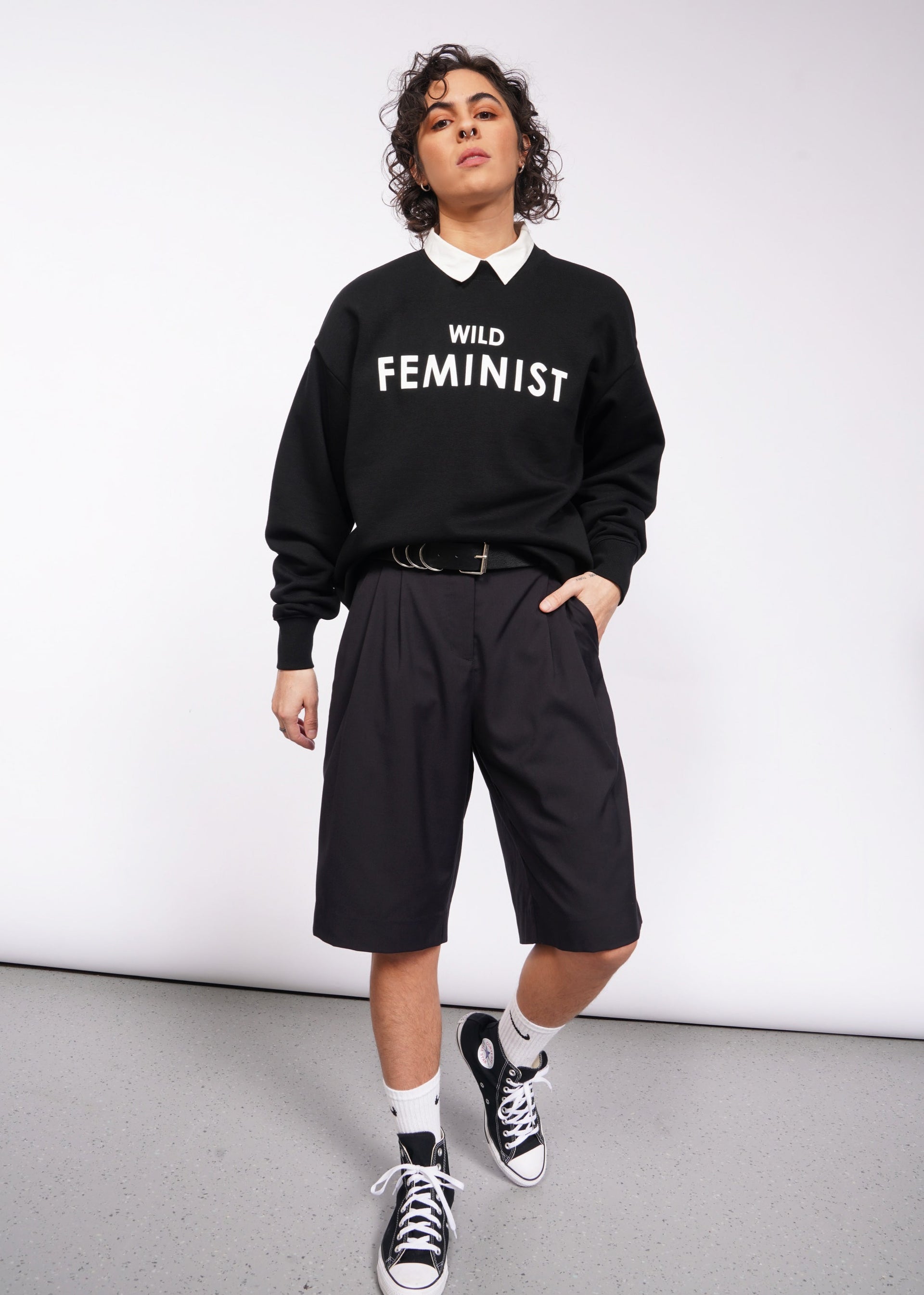 S=A person with short curly hair is wearing the Wild Feminist Fleece Crew over a white collared shirt, paired with black shorts, white socks, and black sneakers. They pose against a plain white background.