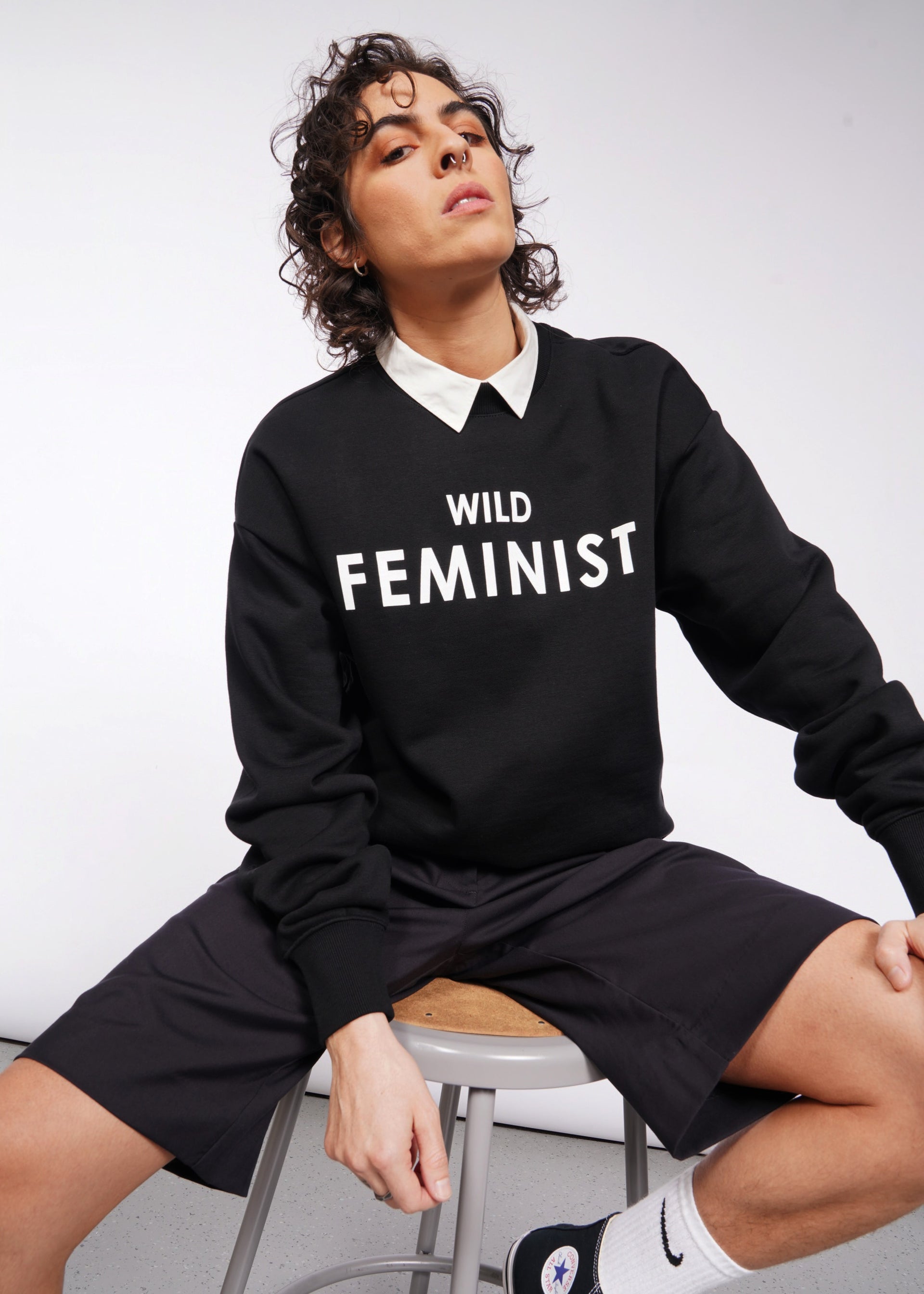 S=A person with curly hair confidently sits on a stool in a minimalist setting, wearing the Wild Feminist Fleece Crew. Their black shorts, white socks, and black shoes perfectly enhance the bold crewneck design.