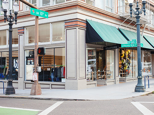 Picture of street corner in Portland with a street sign that says SW 10th Ave. Past the street and sidewalk you see the corner the Wildfang storefront featuring full length glass windows, green awnings, a Wildfang logo decal in the window, and a Wildfang sign hanging from the awning.