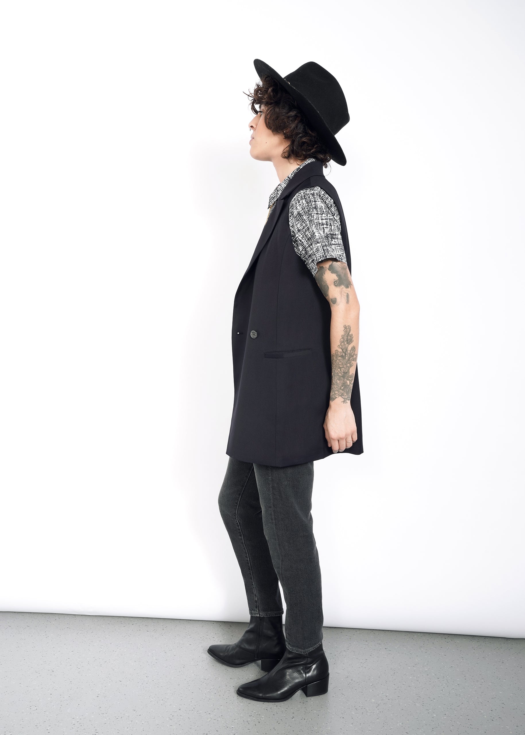 S=With curly hair and tattoos, a person in profile against a white background wears The Empower Double Breasted Vest in an oversized fit, paired with a patterned shirt, black jeans, boots, and a black hat while gazing upwards.