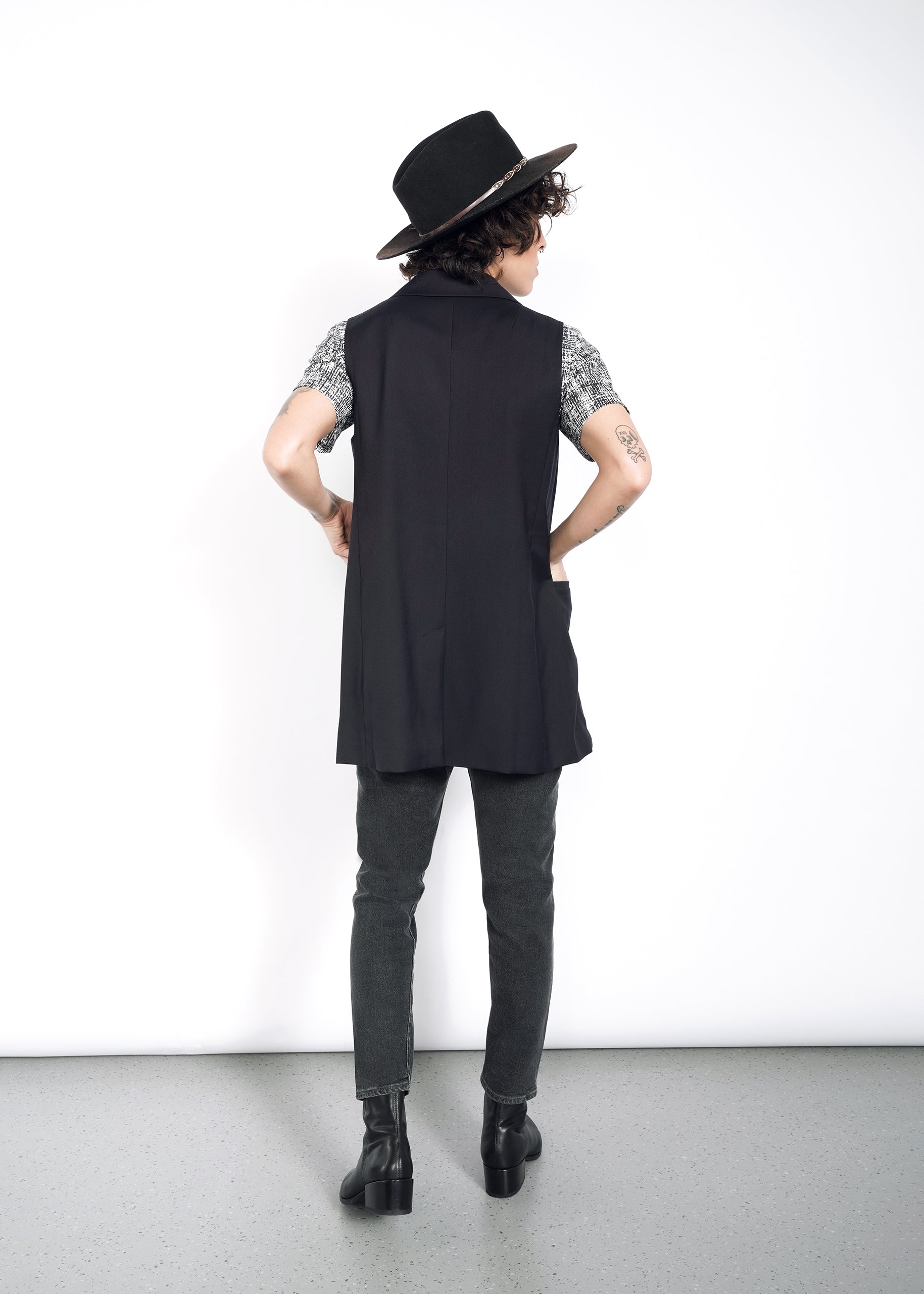 S=A person with short curly hair, wearing a wide-brimmed hat, patterned shirt, The Empower Double Breasted Vest in oversized fit, gray pants, and black boots faces a white wall with arms akimbo. Tattoos on their arms are visible.