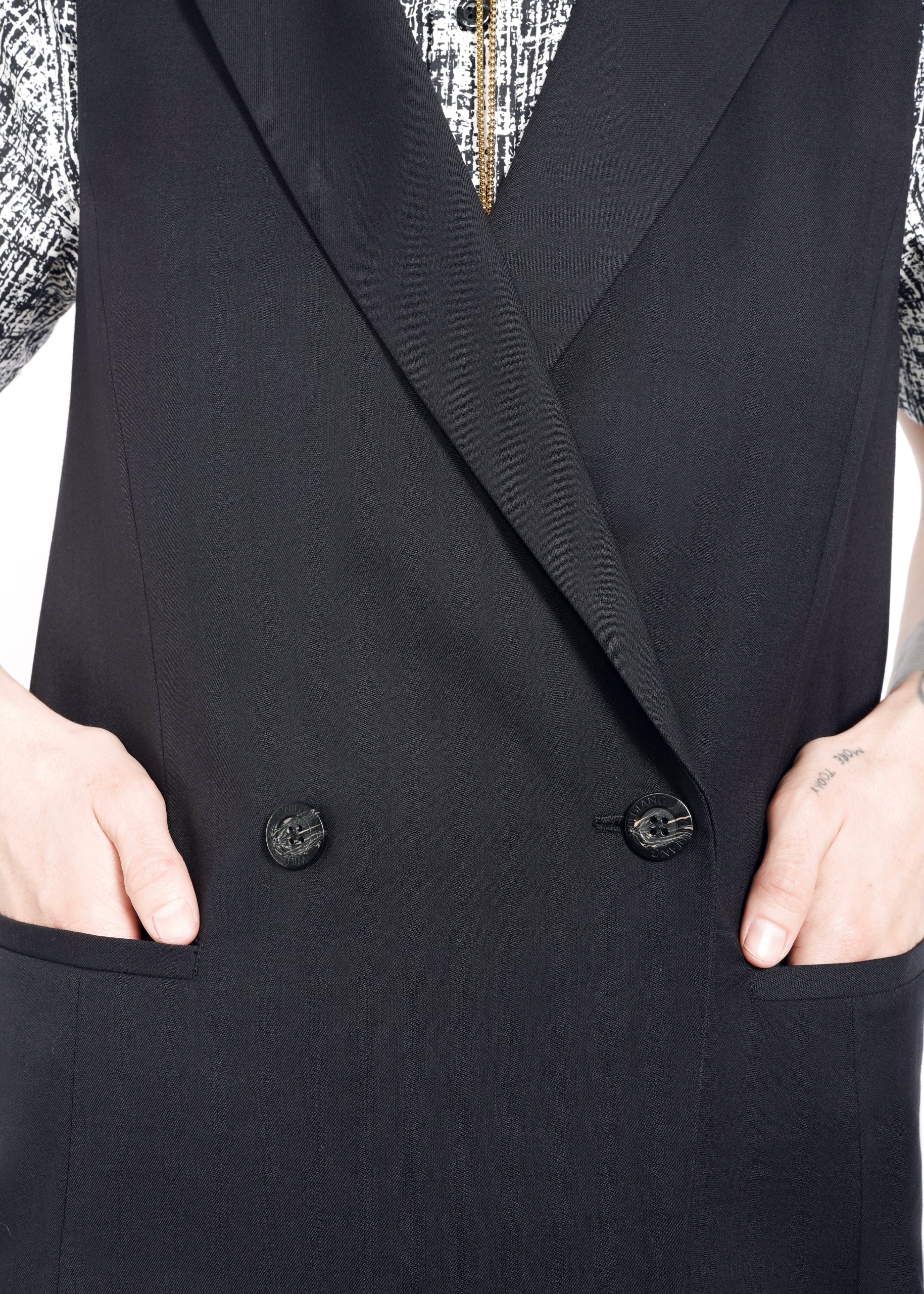 S=A person sporting an oversized fit with The Empower Double Breasted Vest nestles their hands in the pockets. The attire features a patterned shirt underneath, with a close-up image highlighting sleek black buttons dominating the midsection.