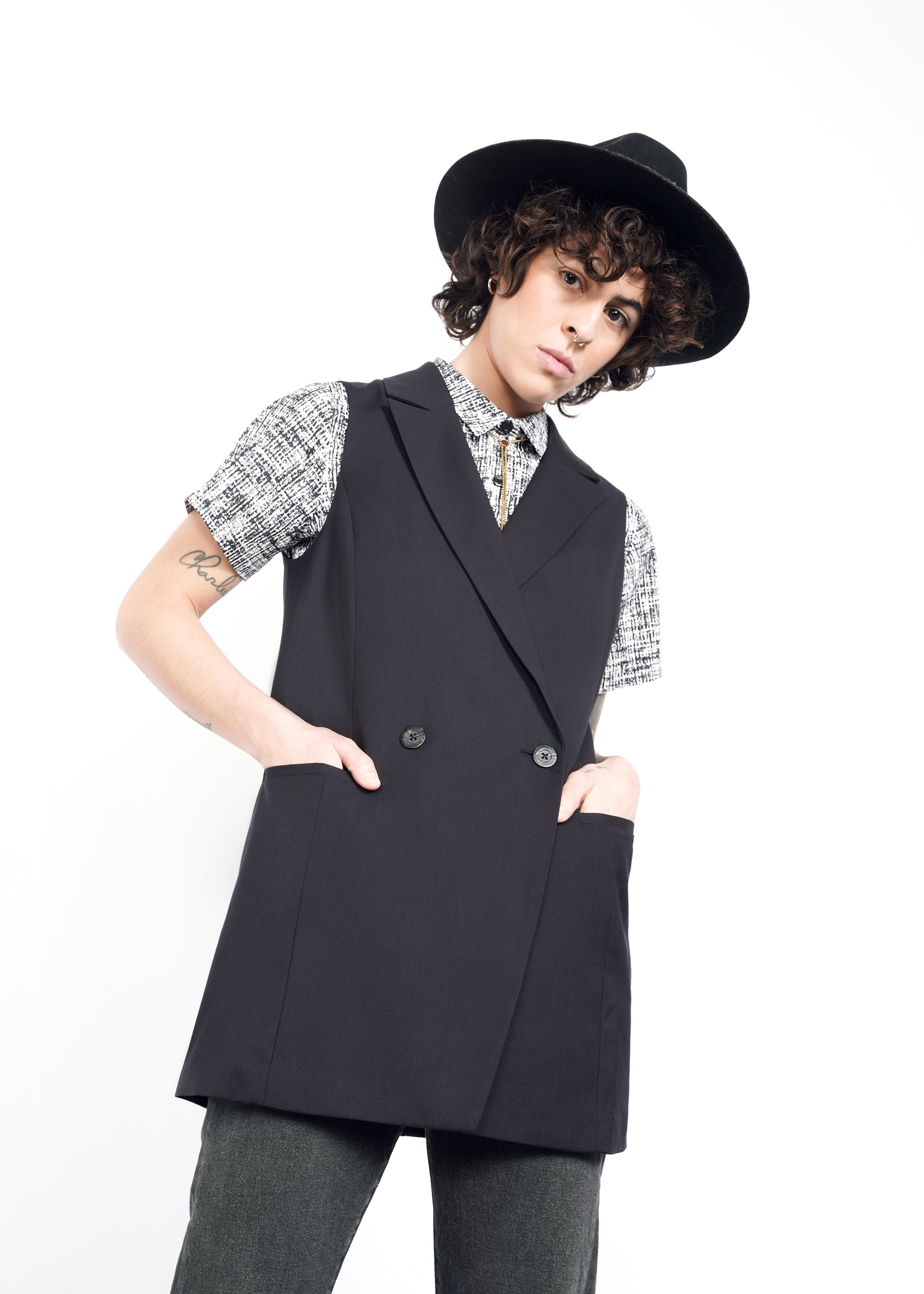 S=A person with short curly hair wears an oversized black wide-brimmed hat, The Empower Double Breasted Vest over a patterned shirt, and gray pants. They stand with hands in pockets against a white background, looking forward with a neutral expression.