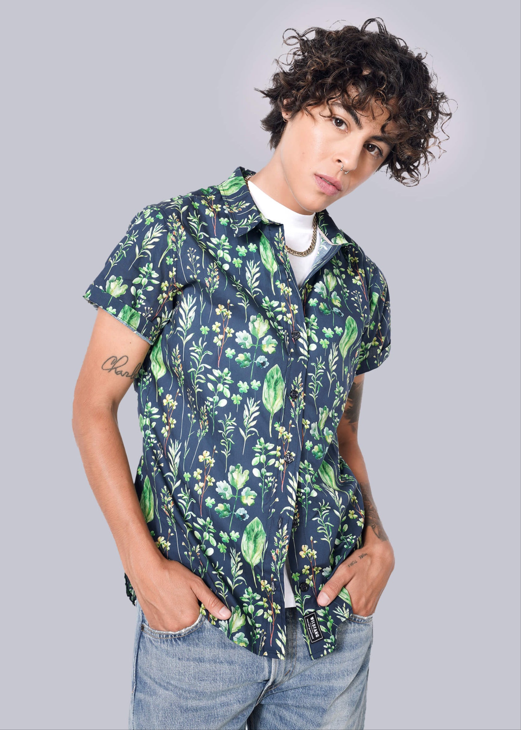 A person with curly hair confidently wears The Essential Button Up in navy with green floral patterns, tailored fit, and short sleeves. Underneath is a white shirt, paired with light blue jeans against a crisp plain white background. Hands are casually tucked in pockets.