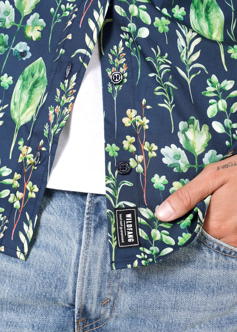 A person is wearing The Essential Button Up—a dark blue, short-sleeved 100% cotton shirt with a green leaf and botanical print. The shirt is partly buttoned over a white top, paired with light blue jeans. Their tattooed hands are visible. A clothing label reads WILDLIING.