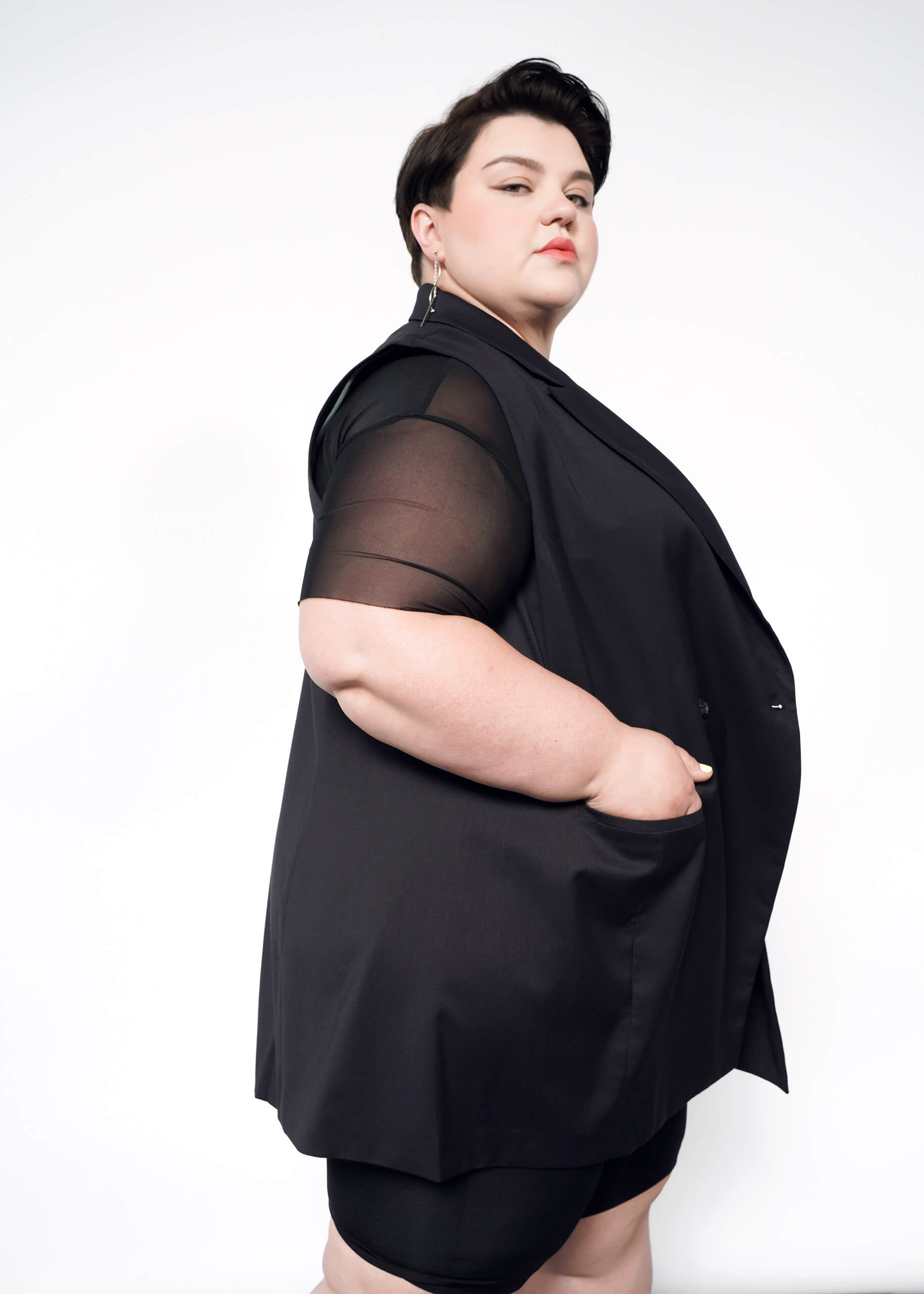 1X=A person with short dark hair stands confidently against a plain white background, wearing The Empower Double Breasted Vest with sheer-sleeved black attire. Their hands are in the pockets, one side of the vest slightly draped open, exuding effortless style.