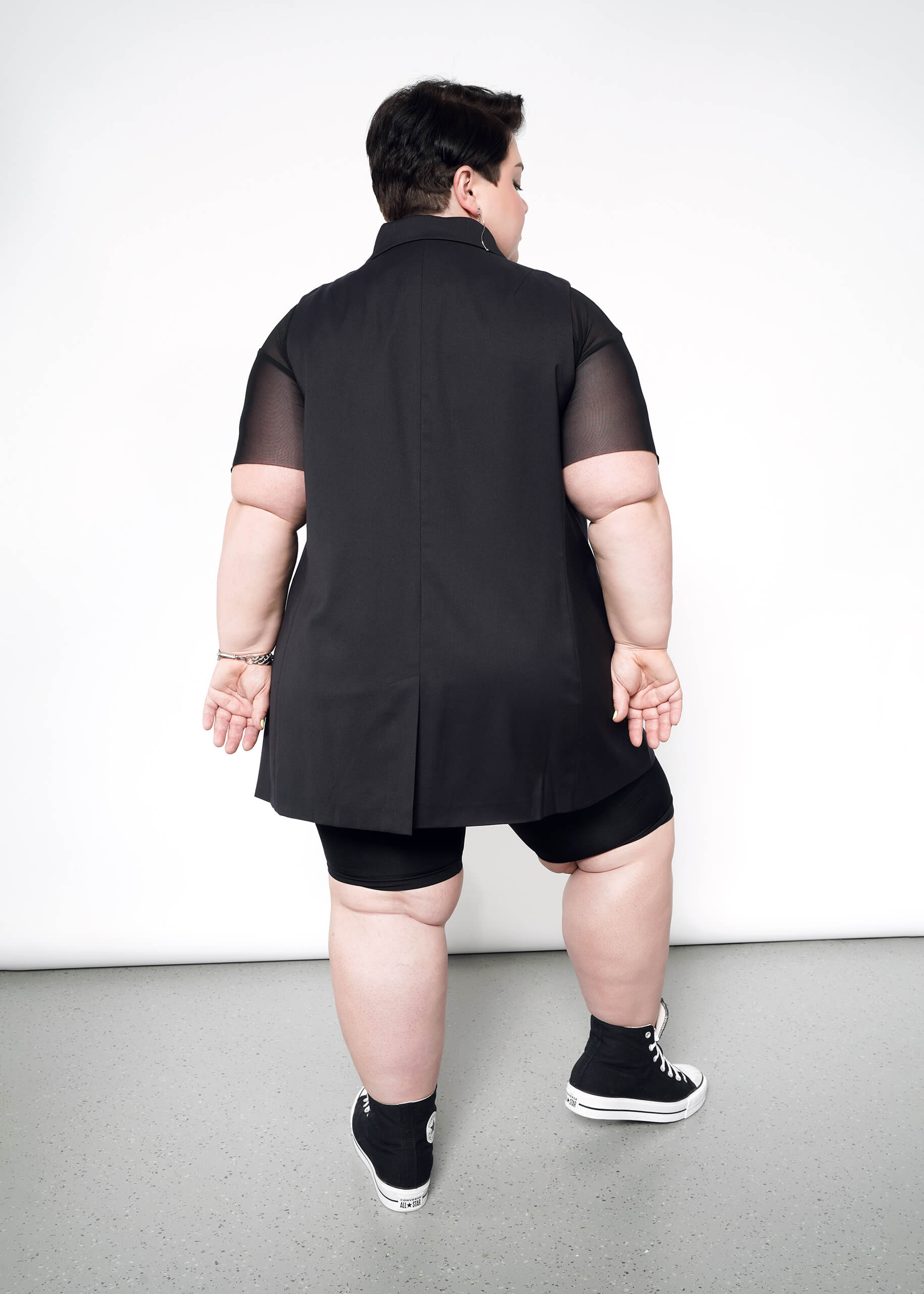 1X=A person stands facing a white wall, wearing The Empower Double Breasted Vest with black short-sleeve shirt, black shorts, and high-top sneakers. Their hands are clasped behind their back.