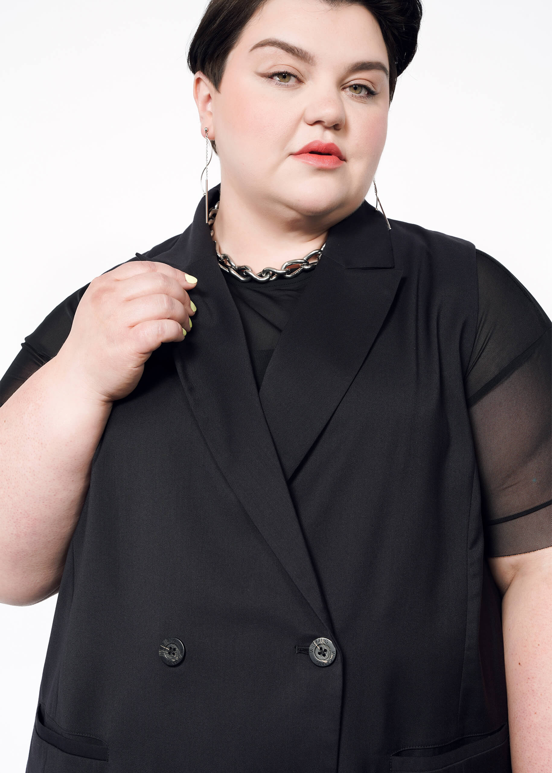 1X=A person with short dark hair wears The Empower Double Breasted Vest, a sleek black sleeveless blazer with an oversized fit, over a mesh top and delicately holds the lapel. Adorned with hoop earrings, a chunky chain necklace, and light pink lipstick, they stand against a crisp white background.