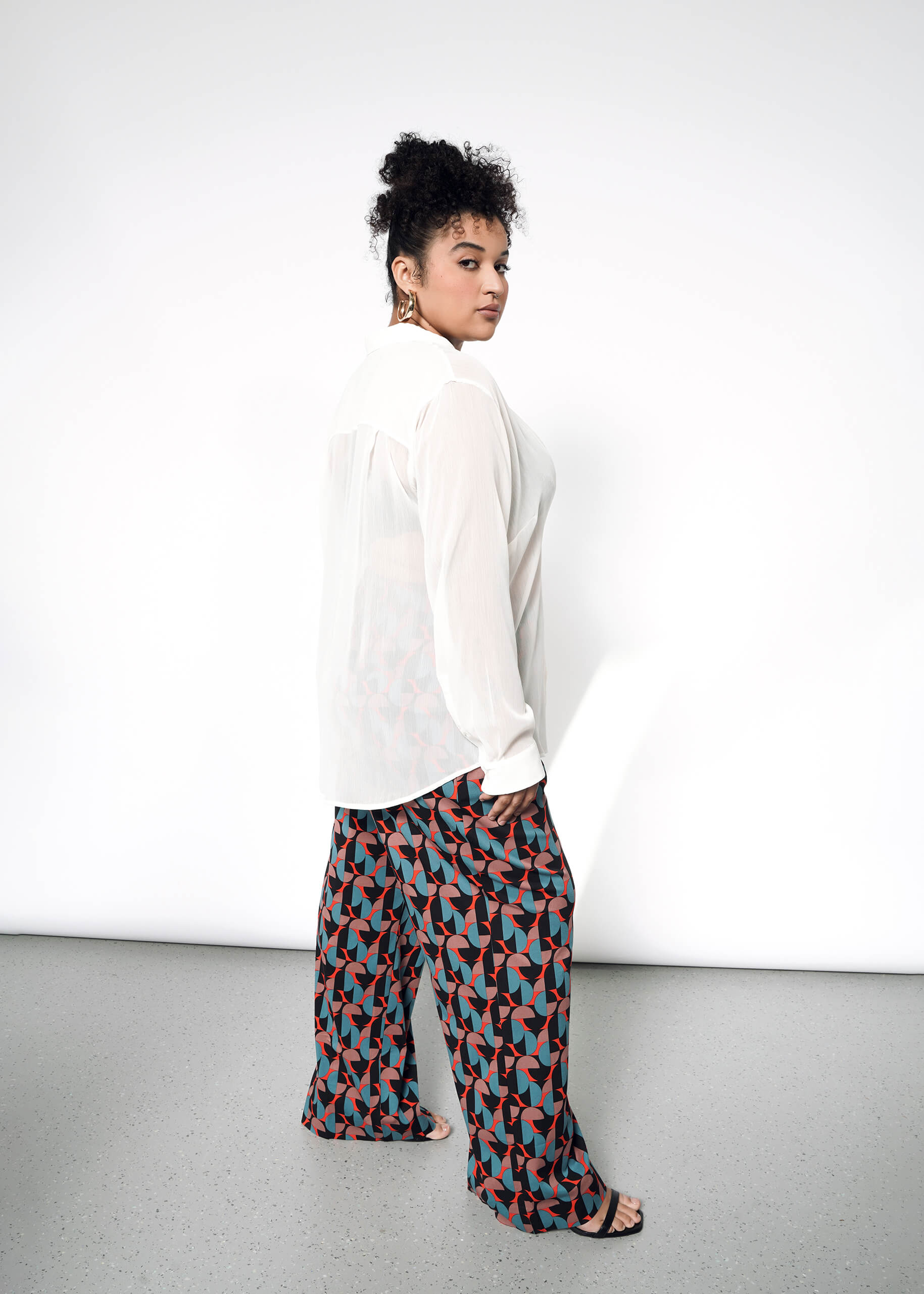 1X=A woman with curly hair models The Empower Sheer Long Sleeve Button Up and wide-leg pants with red, black, and teal geometric patterns, standing sideways against a plain white background.