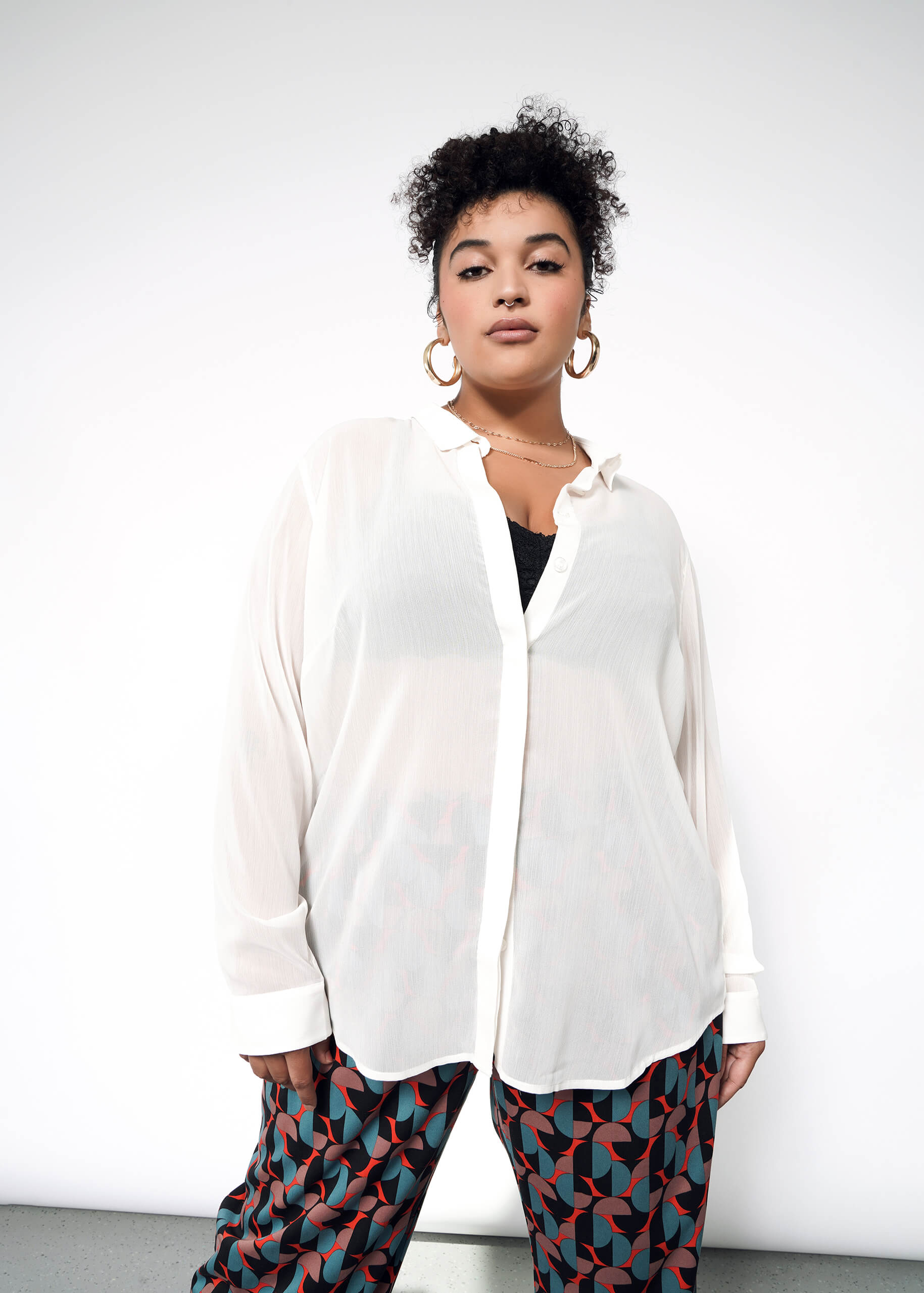 1X= A woman with short curly hair wears The Empower Sheer Long Sleeve Button Up, featuring an oversized fit and Wildfang branded buttons, layered over a black top and patterned pants, standing confidently against a plain white background.