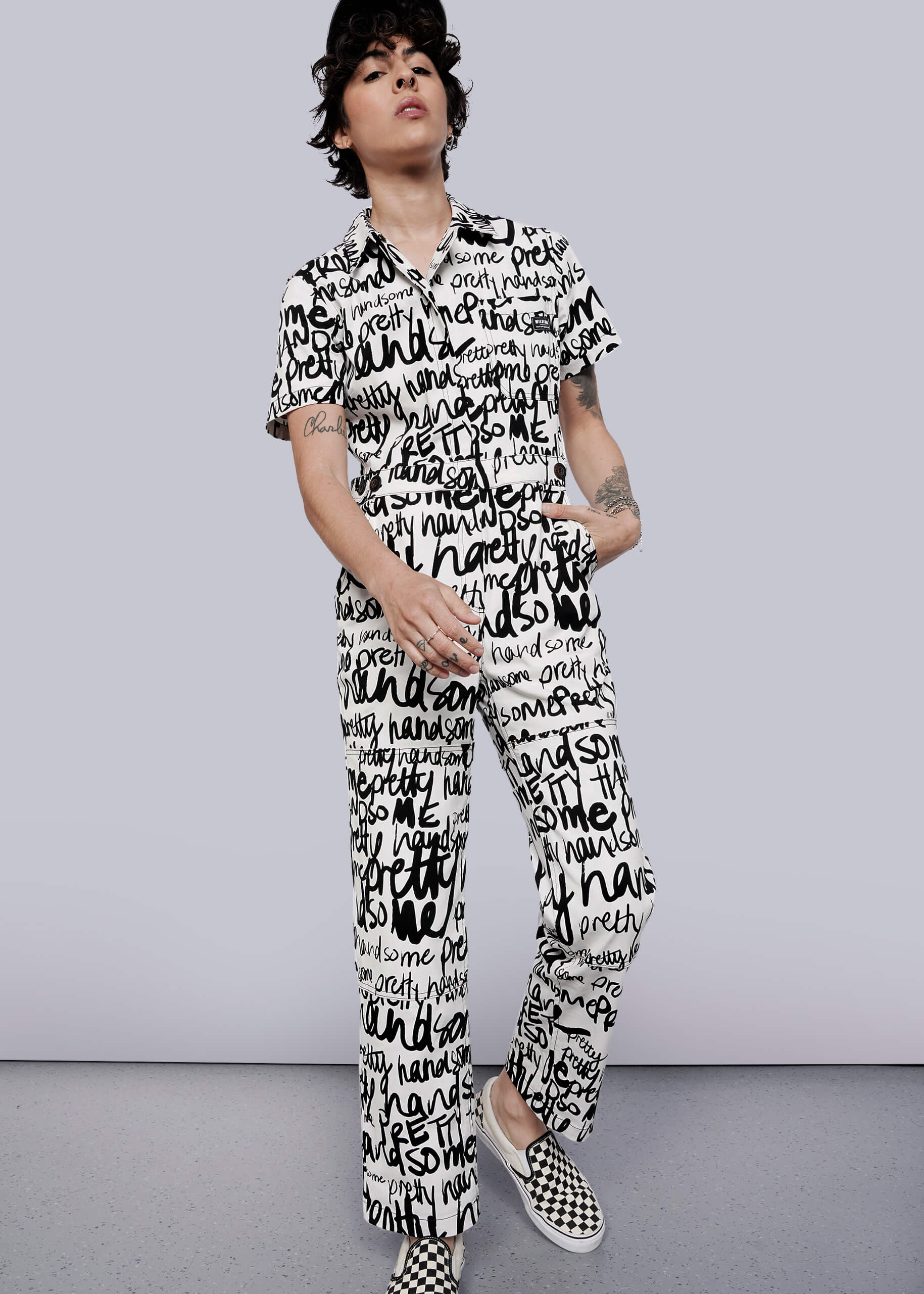 S=A person with short dark hair and arm tattoos wears The Essential High Waisted Coverall—a white jumpsuit with black handwritten words—paired with black-and-white checkered slip-on shoes, standing against a plain white background.
