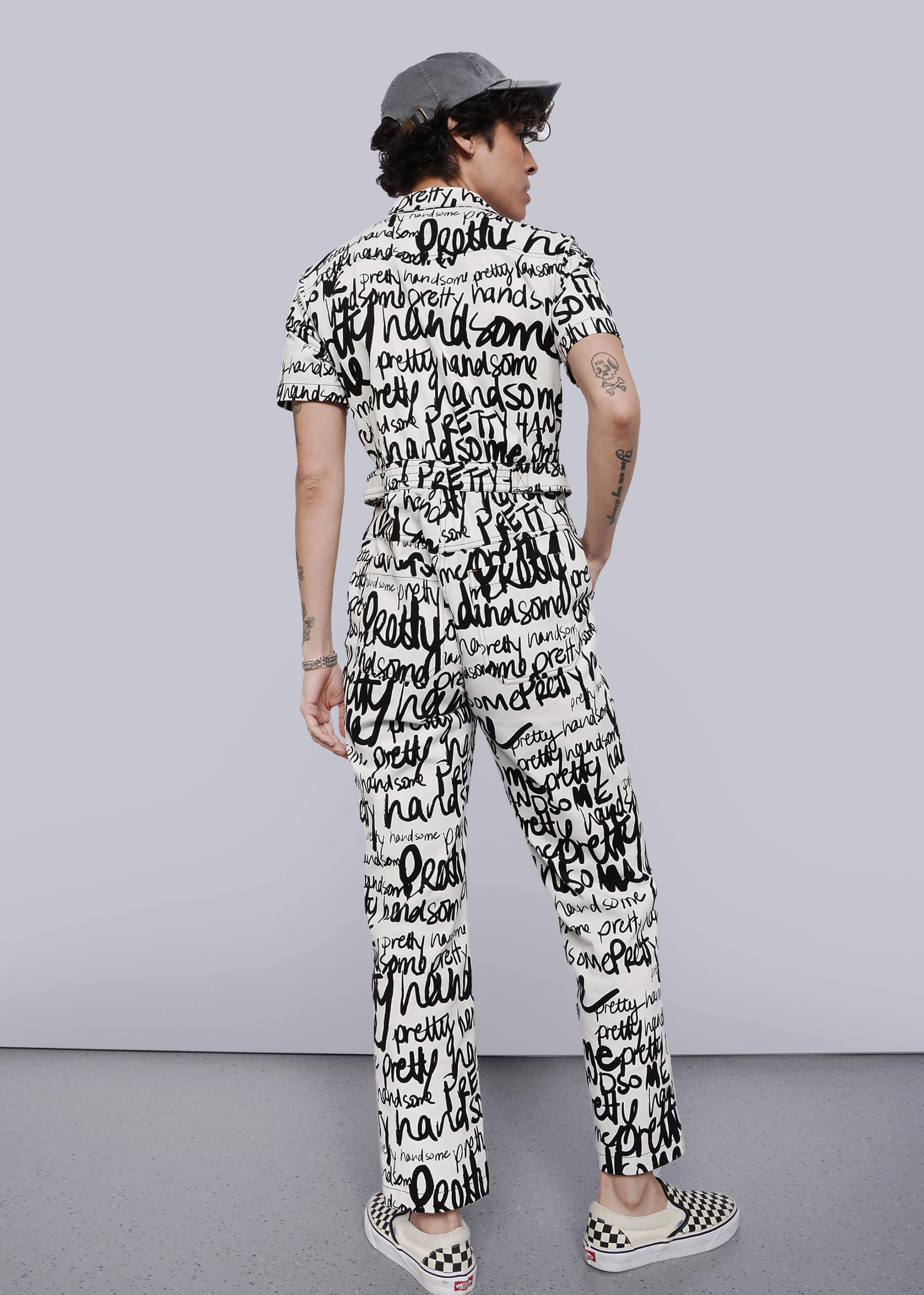 S=A person stands facing away, wearing The Essential High Waisted Coverall—a black-and-white jumpsuit with handwritten words—plus black-and-white checkerboard slip-on shoes and a gray cap. The background is plain and light-colored.