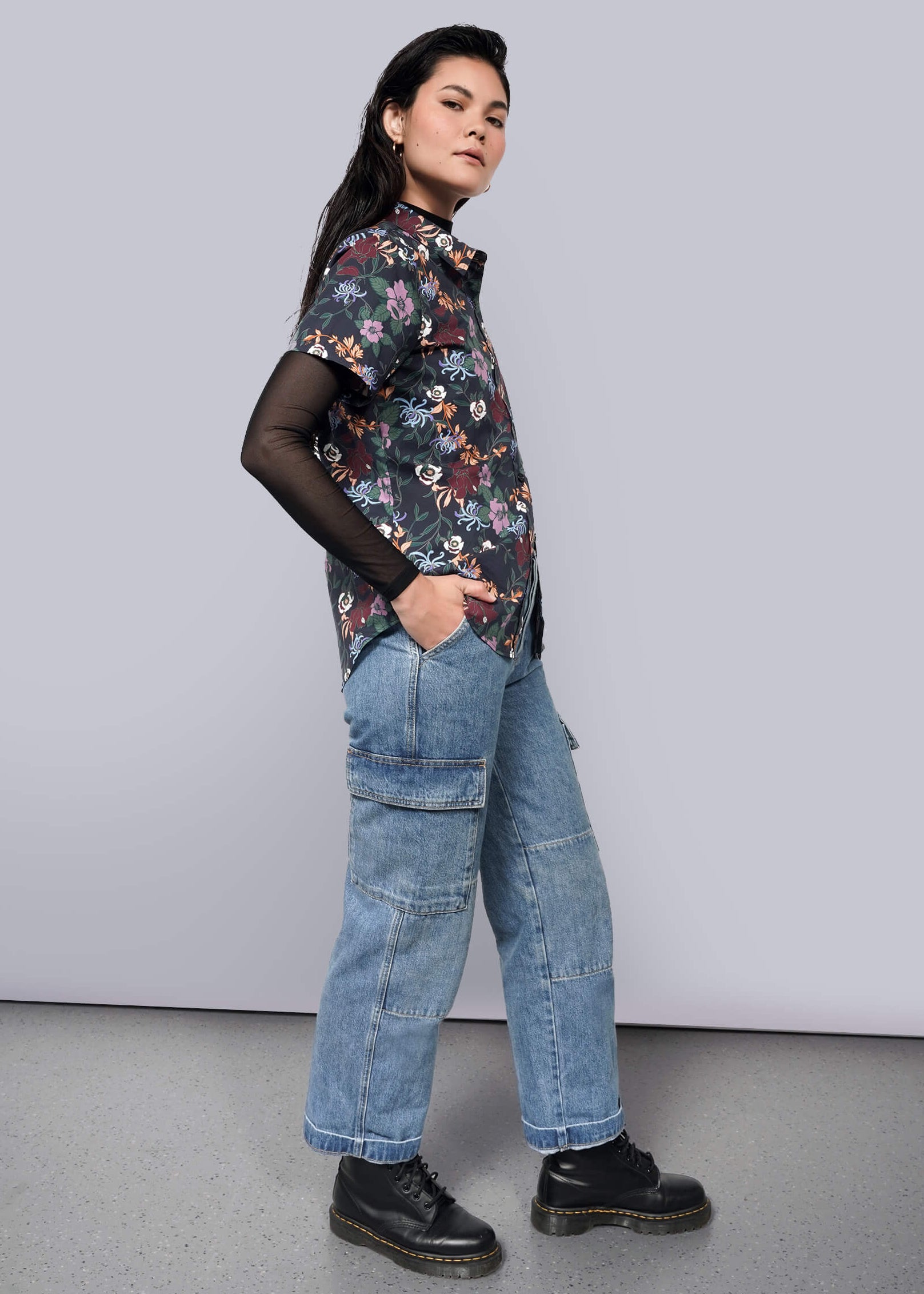 S=A woman stands confidently against a plain background, wearing The Essential Button Up by Wildfang over a sheer black top, wide-leg blue jeans with cargo pockets, and black platform shoes. She completes her tailored look with hands in her pockets.