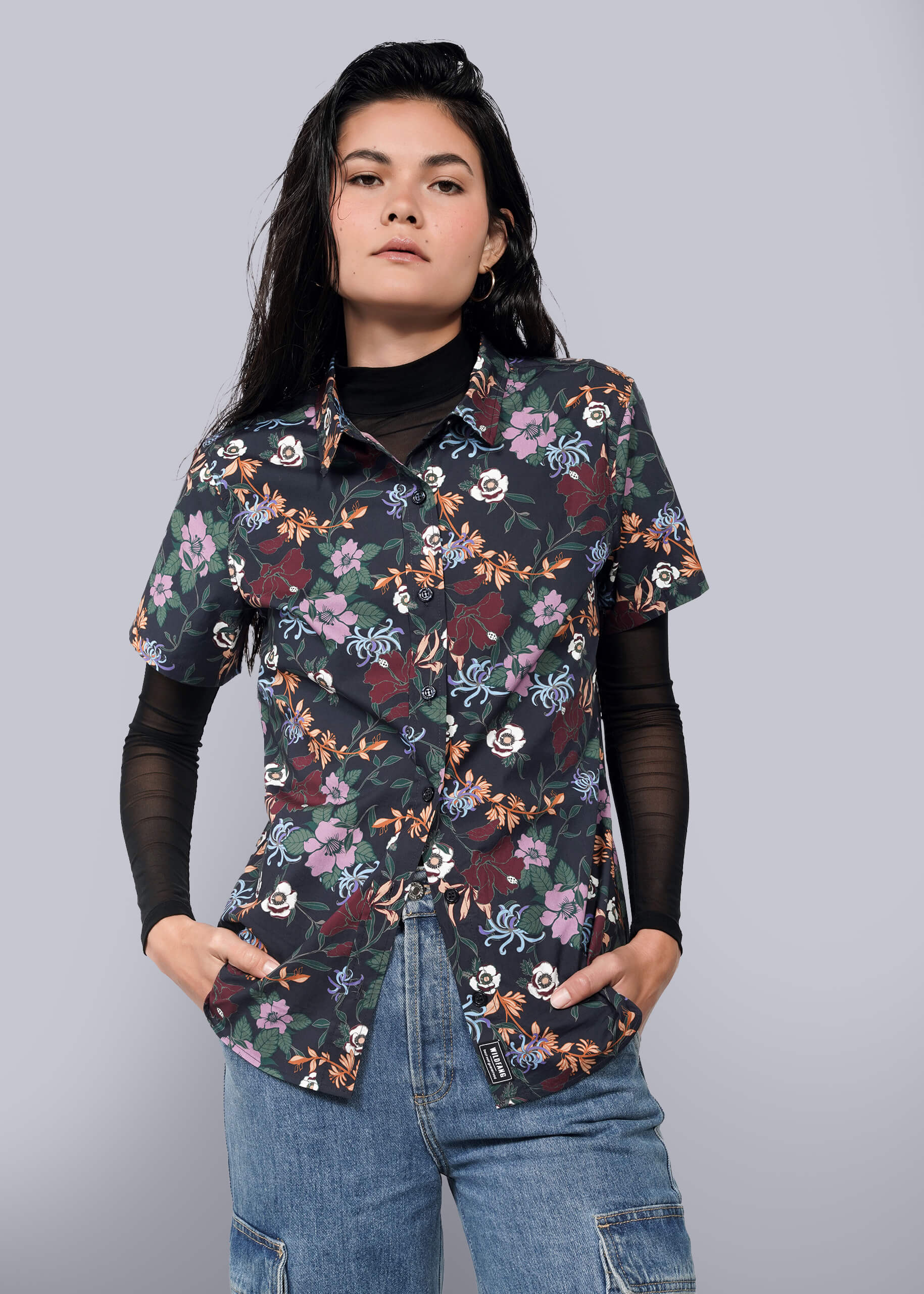 S= A person with long dark hair wears The Essential Button Up in black print, styled over a black long-sleeve top and blue jeans, standing with hands in pockets against a plain white background.