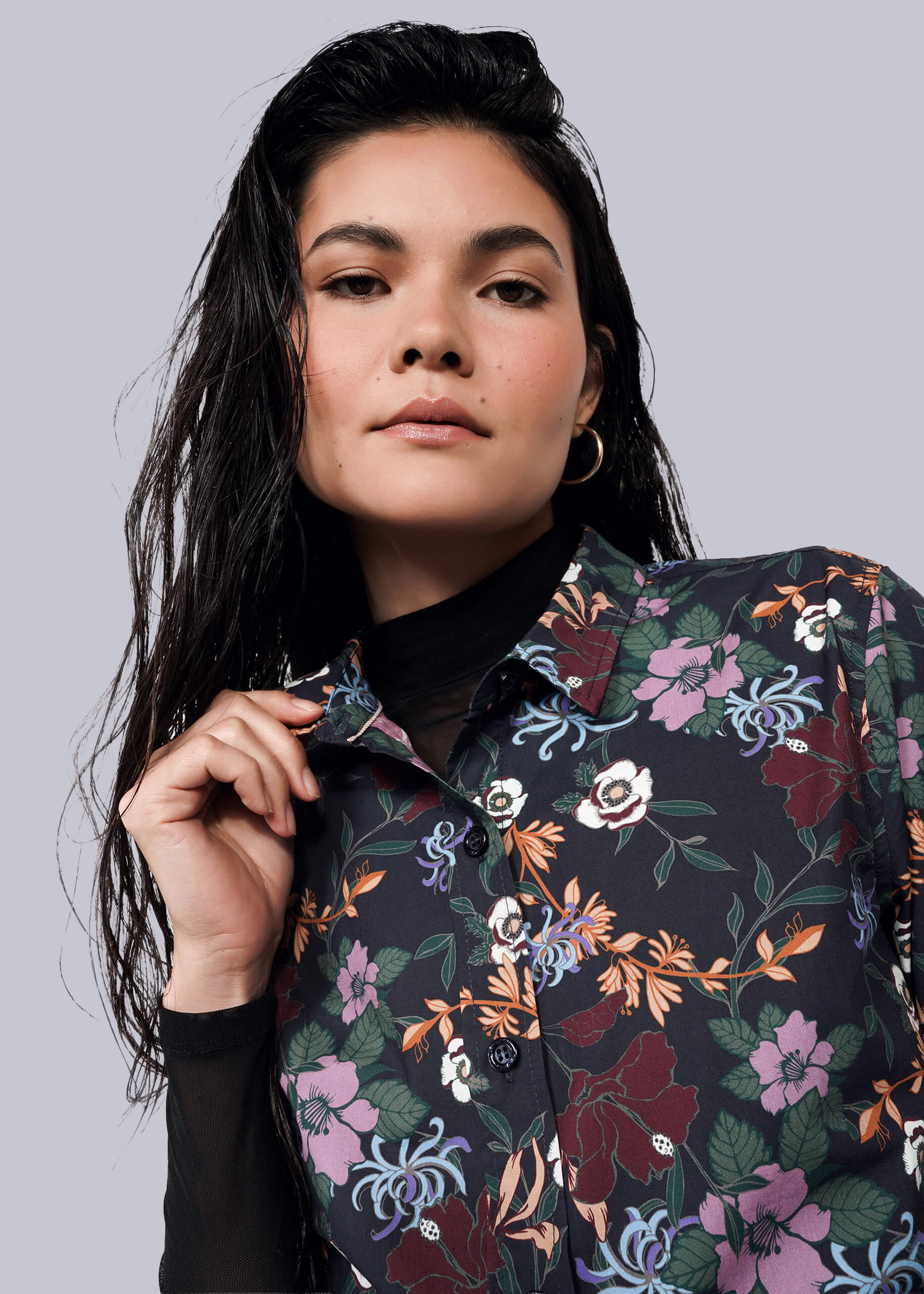 S=A woman with long dark hair wears The Essential Button Up—a tailored, floral-patterned shirt—over a black top. She confidently holds the collar, no chest gap visible, while looking at the camera against a plain white background.