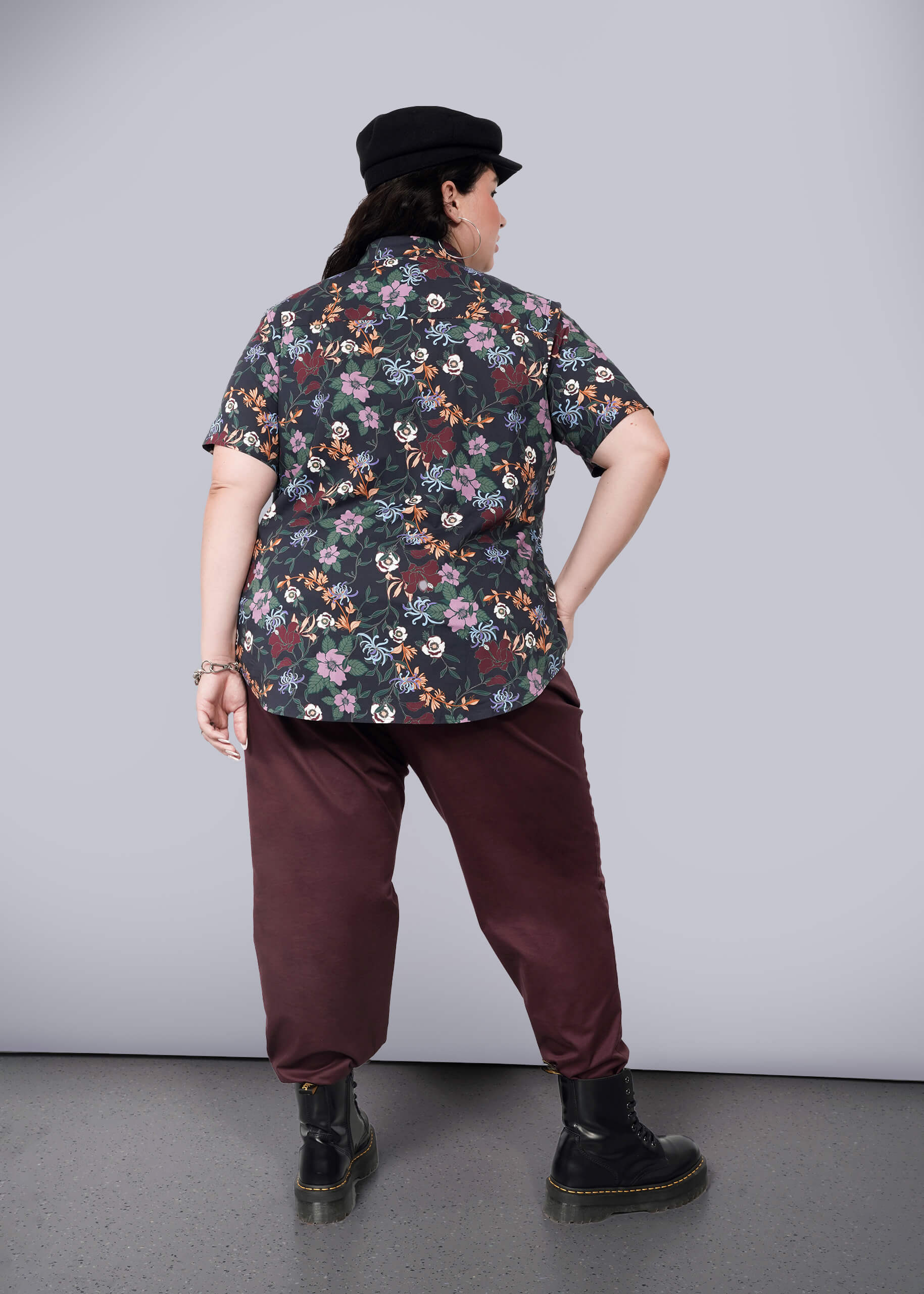 1X= A person faces away, wearing a black cap, The Essential Button Up in dark Wildfang floral print with a tailored fit and no chest gap, burgundy pants, and black boots, one hand on their hip against a plain white background.