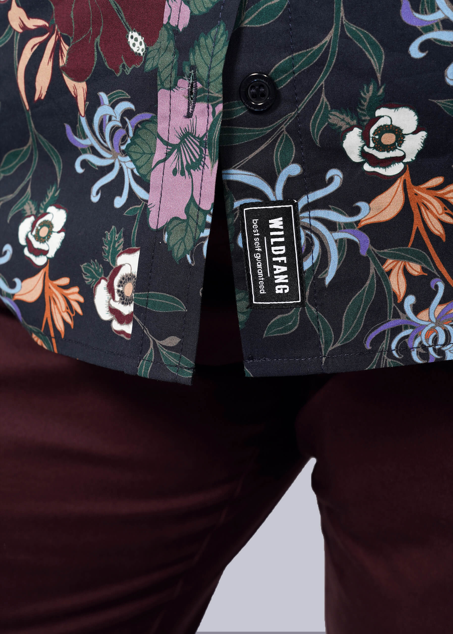 1X=Close-up of a person wearing maroon pants and The Essential Button Up, featuring a vibrant Wildfang print. A WILDFANG brand tag is visible by the hem, showcasing the shirt’s tailored fit and attention to detail.